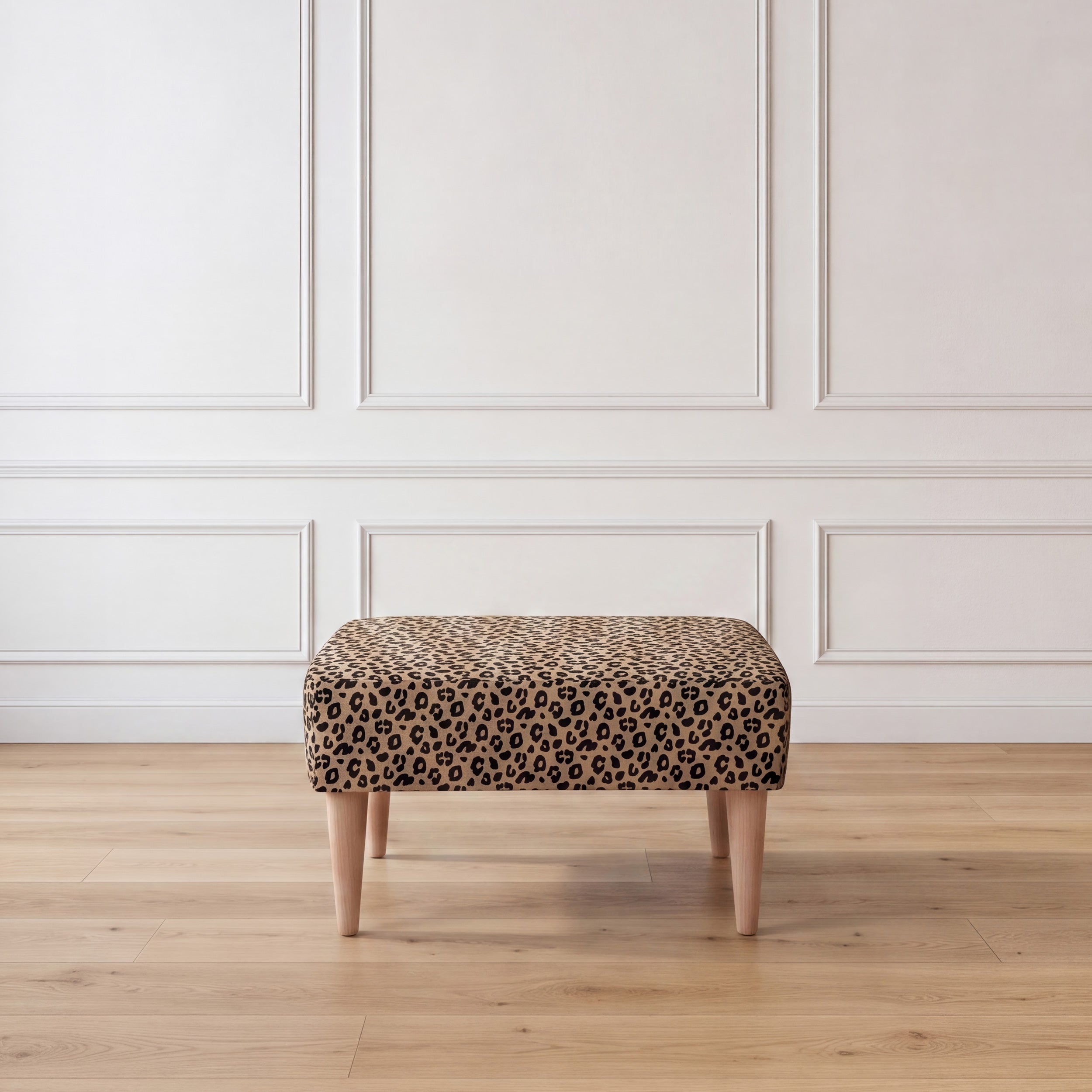 A rectangle upholstered ottoman with Sahara Leopard print pattern supported by four light wood legs, set against a white paneled wall and light wood floor.