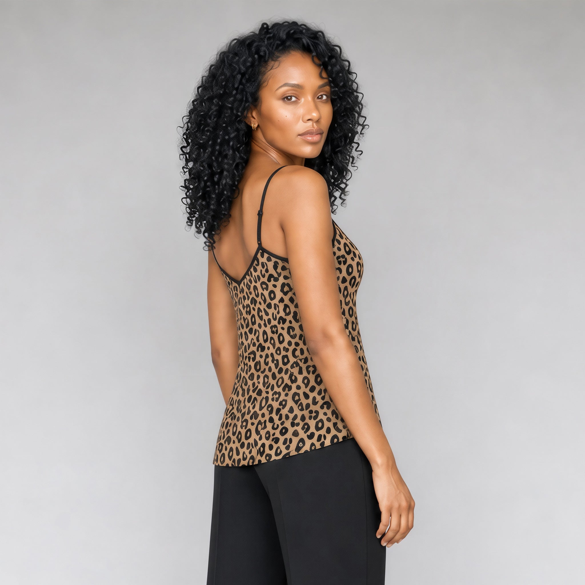 A 45 degree angle back view of a black woman with curly black hair wearing a Sahara leopard print camisole top with black trim, paired with black pants, standing against a neutral gray studio background.