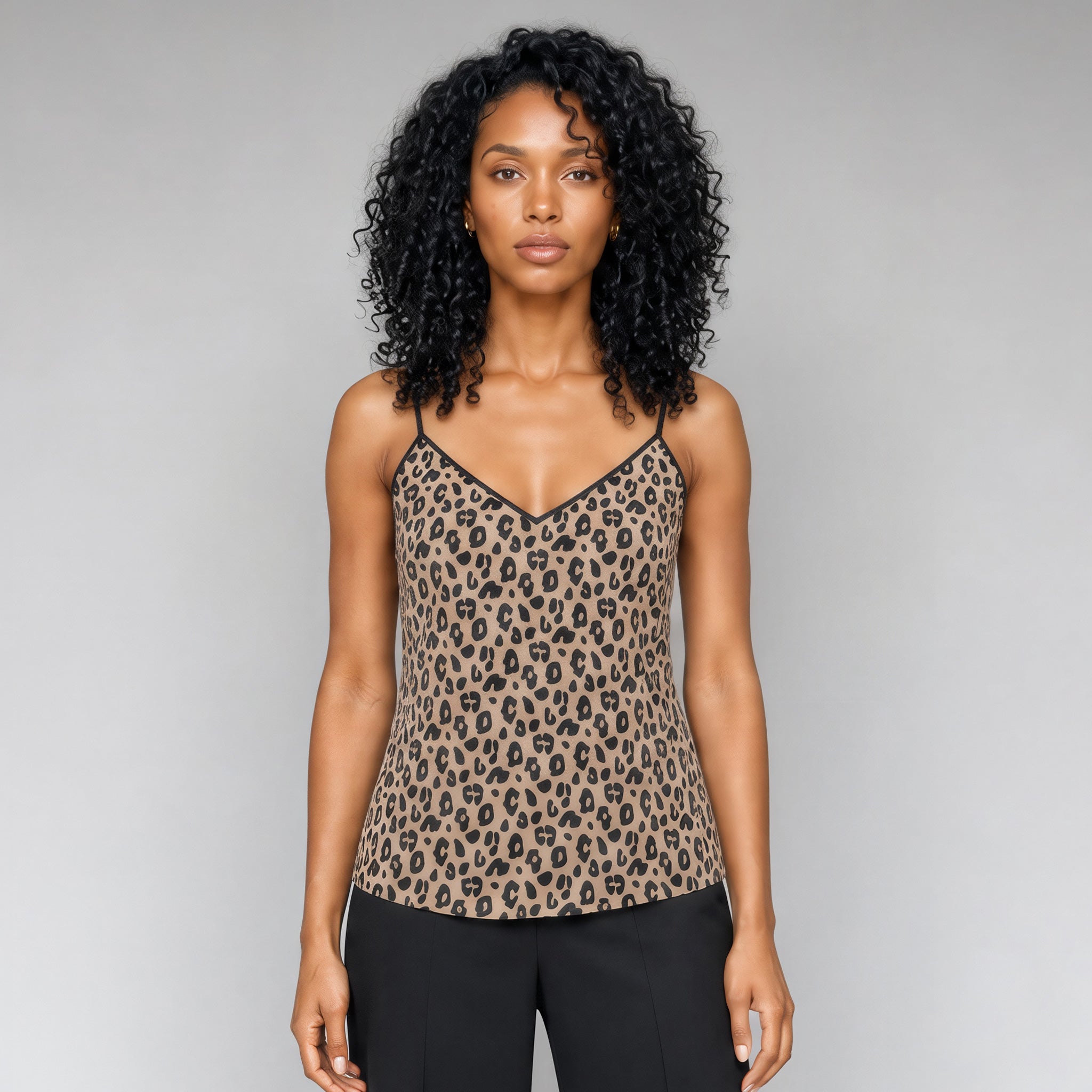 A front view of a black woman with curly black hair wearing a Sahara leopard print camisole top with black trim, paired with black pants, standing against a neutral gray studio background.