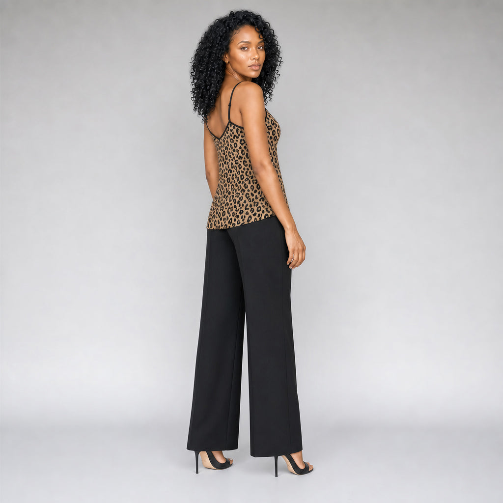 A 45 degree angle full back view of a black woman with curly black hair wearing a Sahara leopard print camisole top with black trim, paired with black pants, and strappy black stilettos, standing against a neutral gray studio background.
