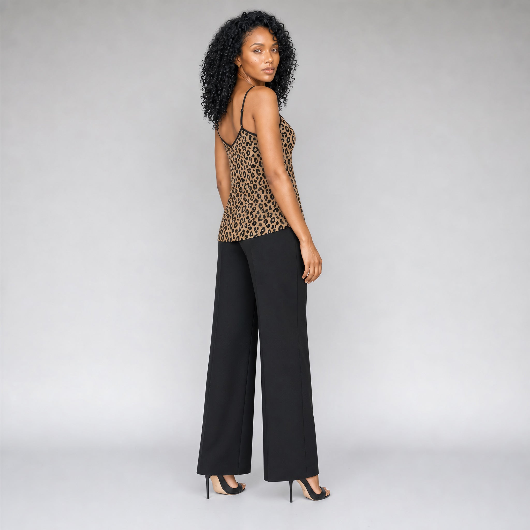 A 45 degree angle full back view of a black woman with curly black hair wearing a Sahara leopard print camisole top with black trim, paired with black pants, and strappy black stilettos, standing against a neutral gray studio background.
