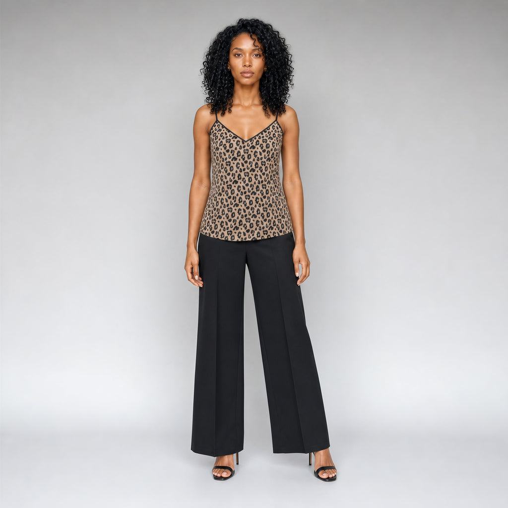 A full front view of a black woman with curly black hair wearing a Sahara leopard print camisole top with black trim, paired with black pants, and strappy black stilettos, standing against a neutral gray studio background.