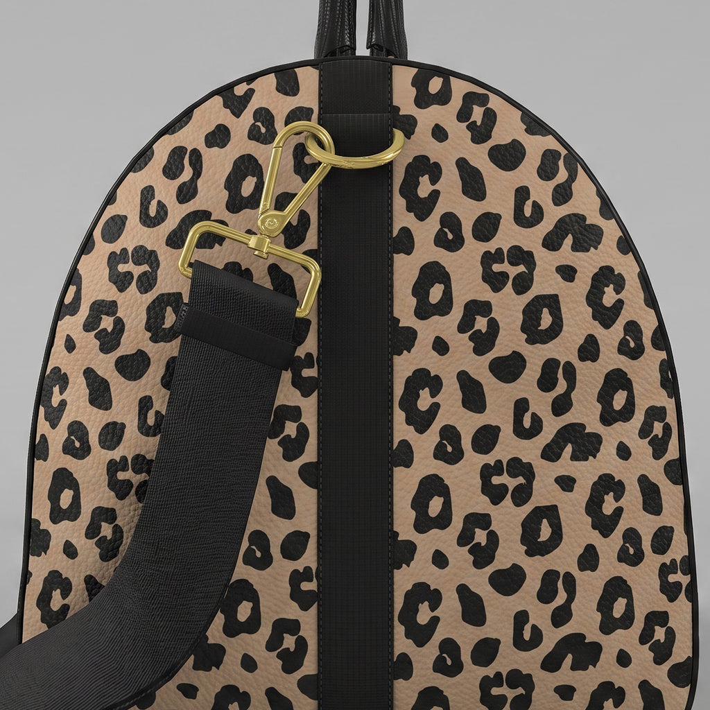 A close-uoleft view of a Sahara leopard print large duffel bag with black trim, a black top handle, and a black shoulder strap with gold-tone hardware, shown against a light gray background.