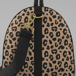 A close-uoleft view of a Sahara leopard print large duffel bag with black trim, a black top handle, and a black shoulder strap with gold-tone hardware, shown against a light gray background.