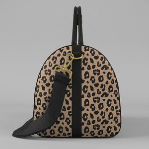 A left view of a Sahara leopard print large duffel bag with black trim, a black top handle, and a black shoulder strap with gold-tone hardware, shown against a light gray background.