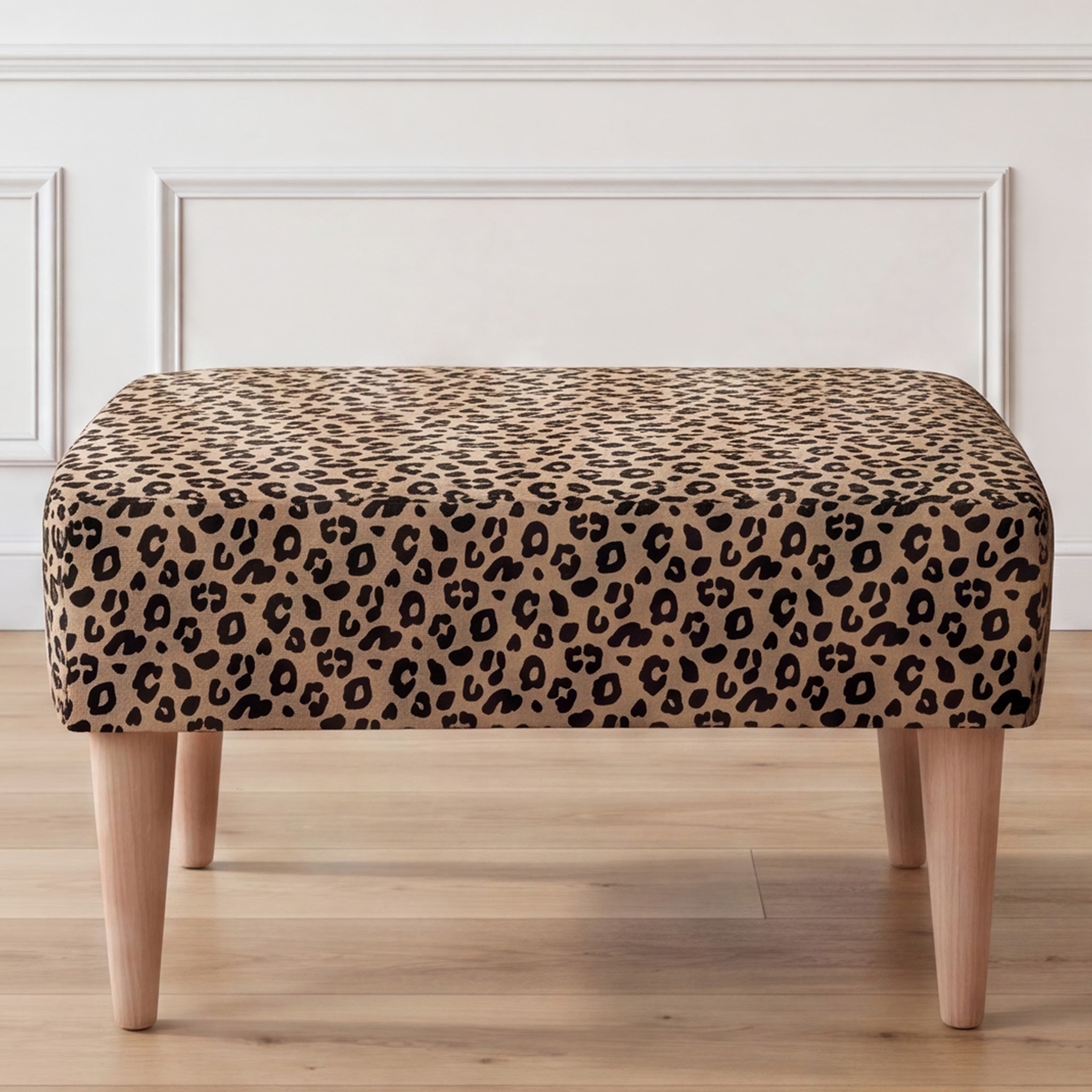 A close-up front view of an ottoman with a leopard print fabric top and four tapered light wood legs, set against a white paneled wall and light wood floor.