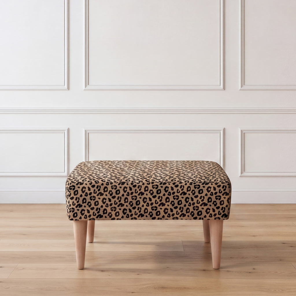 A front view of an ottoman with a leopard print fabric top and four tapered light wood legs, set against a white paneled wall and light wood floor.