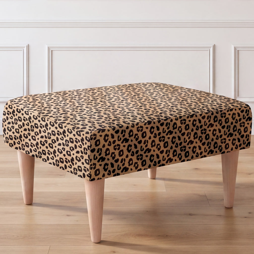 A close-up side view of an ottoman with a leopard print fabric top and four tapered light wood legs, set against a white paneled wall and light wood floor.