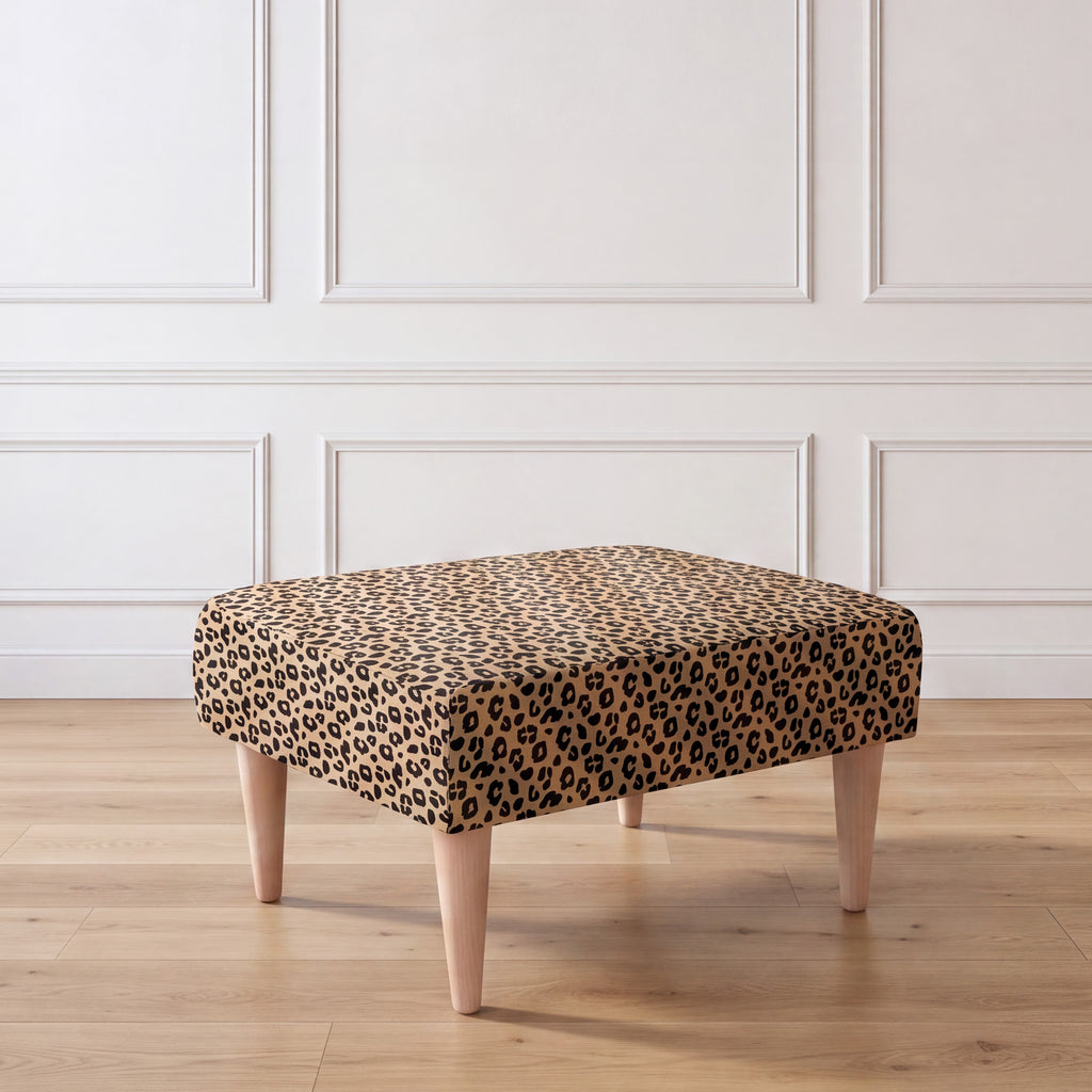 A side view of an ottoman with a leopard print fabric top and four tapered light wood legs, set against a white paneled wall and light wood floor.