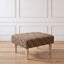 A side view of an ottoman with a leopard print fabric top and four tapered light wood legs, set against a white paneled wall and light wood floor.